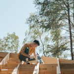 A carpenter works on a wooden roof frame outdoors under clear skies.